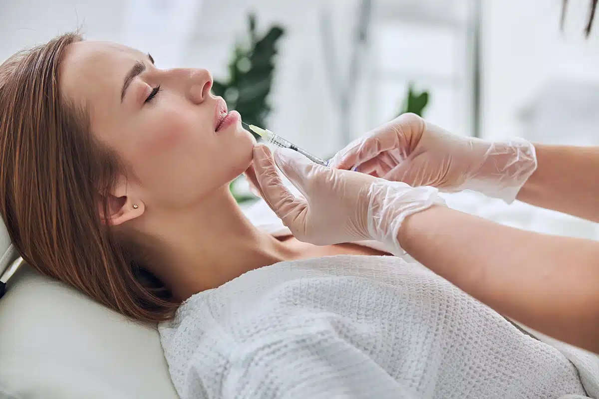 Charming Caucasian young woman with close eyes lying on medicine chair while receiving dermal filler